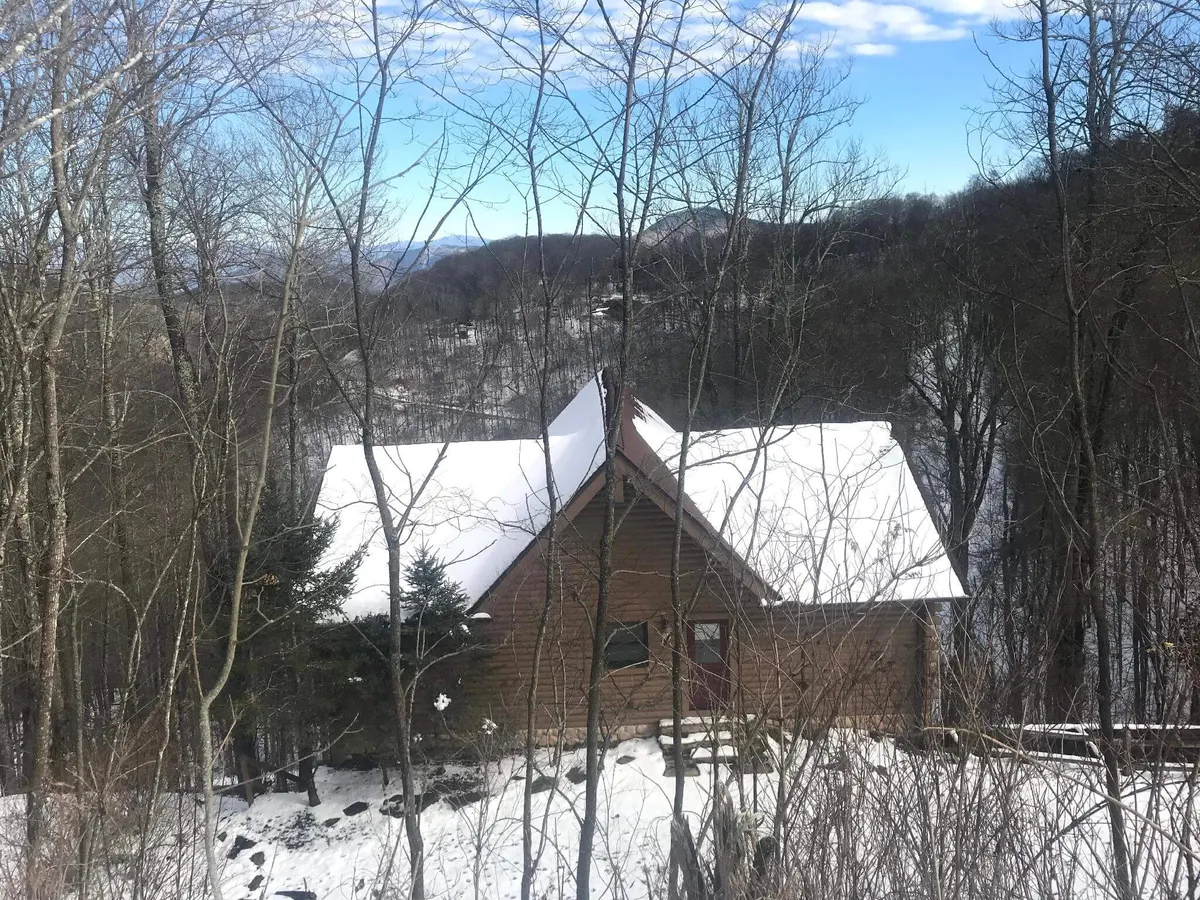 A log cabin with snow on its roof sits nestled among tall trees, with a wide deck visible in the foreground. The scene opens onto a picturesque mountain backdrop, suggesting the home’s elevated setting and natural seclusion.
