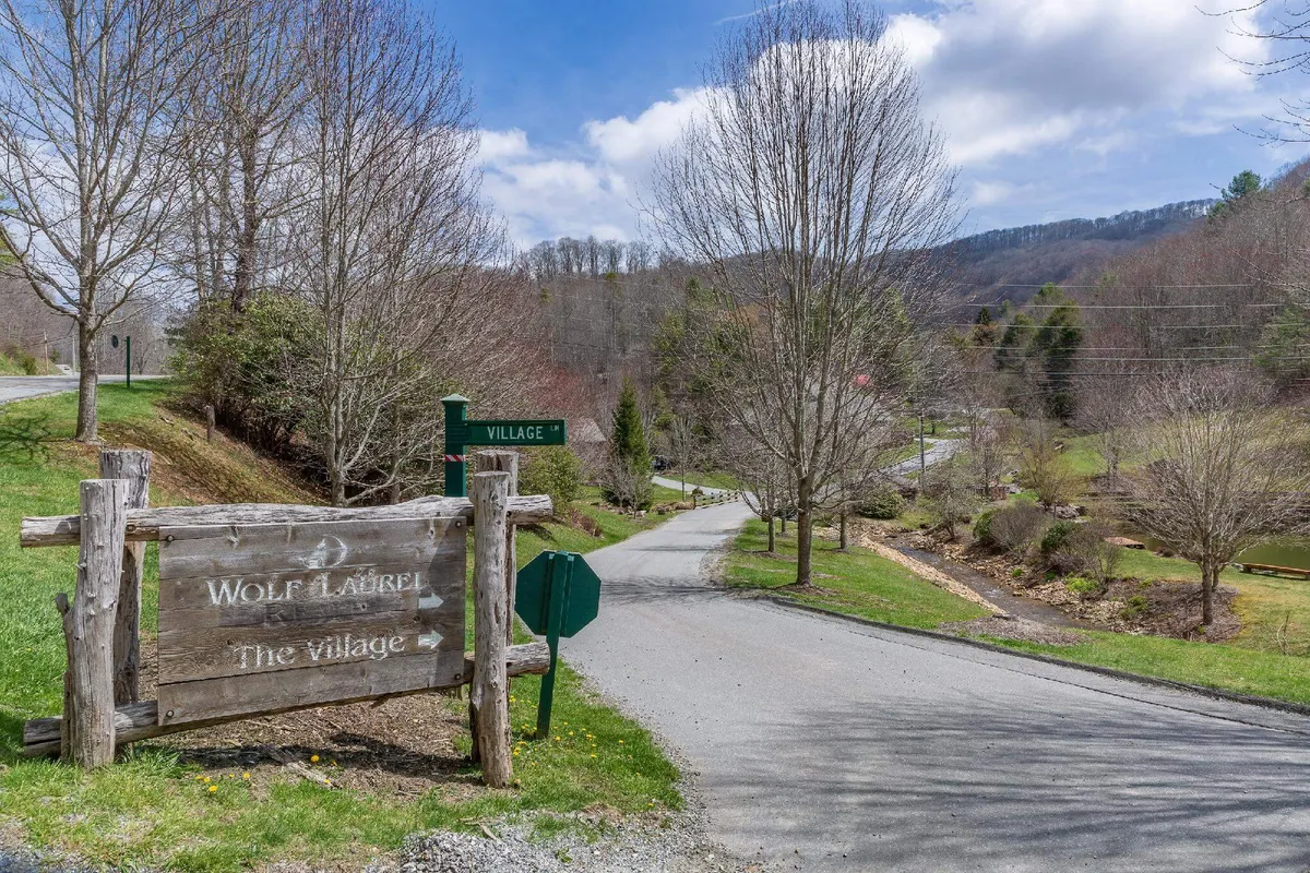 This image captures the welcoming entrance to Wolf Laurel, with a wooden sign reading 'Wolf Laurel' and 'The Village,' a road meandering into the community, and lush trees framing the scene. The inviting landscape suggests both privacy and access to communal resort amenities.