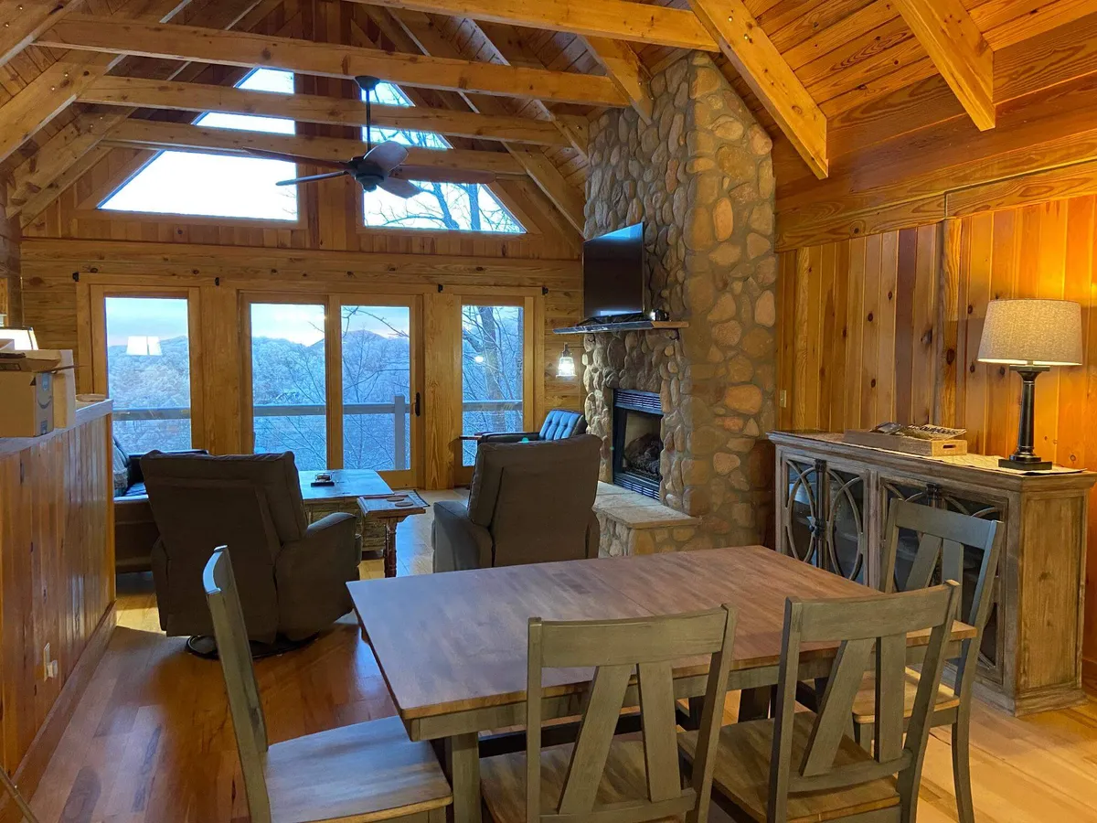 A warm, inviting living and dining area with large windows that frame breathtaking mountain views. The space features a stone fireplace, a spacious dining table, and a comfortable seating area—all bathed in natural light. The arrangement encourages family gatherings, cozy evenings by the fire, and enjoying meals surrounded by nature.