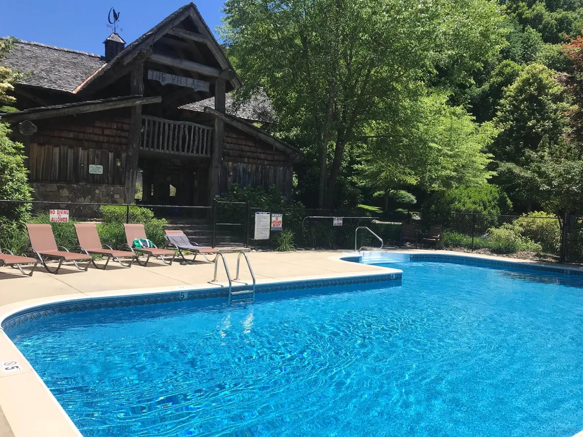 A wooden lodge with two levels is flanked by a sparkling swimming pool, surrounded by comfortable lounge chairs and lush greenery. The decks on both levels are visible and inviting, offering multiple vantage points for outdoor relaxation and gatherings.