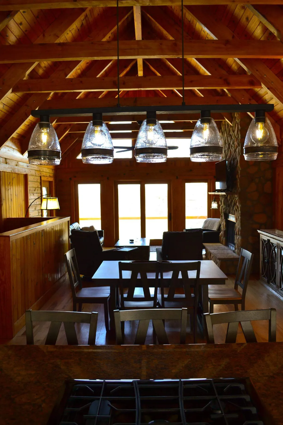 A beautifully appointed open-concept kitchen and dining space with a large wooden dining table, comfortable seating, a stone fireplace, and prominent large windows. The atmosphere is both welcoming and elegant, perfect for socializing or enjoying a quiet evening.