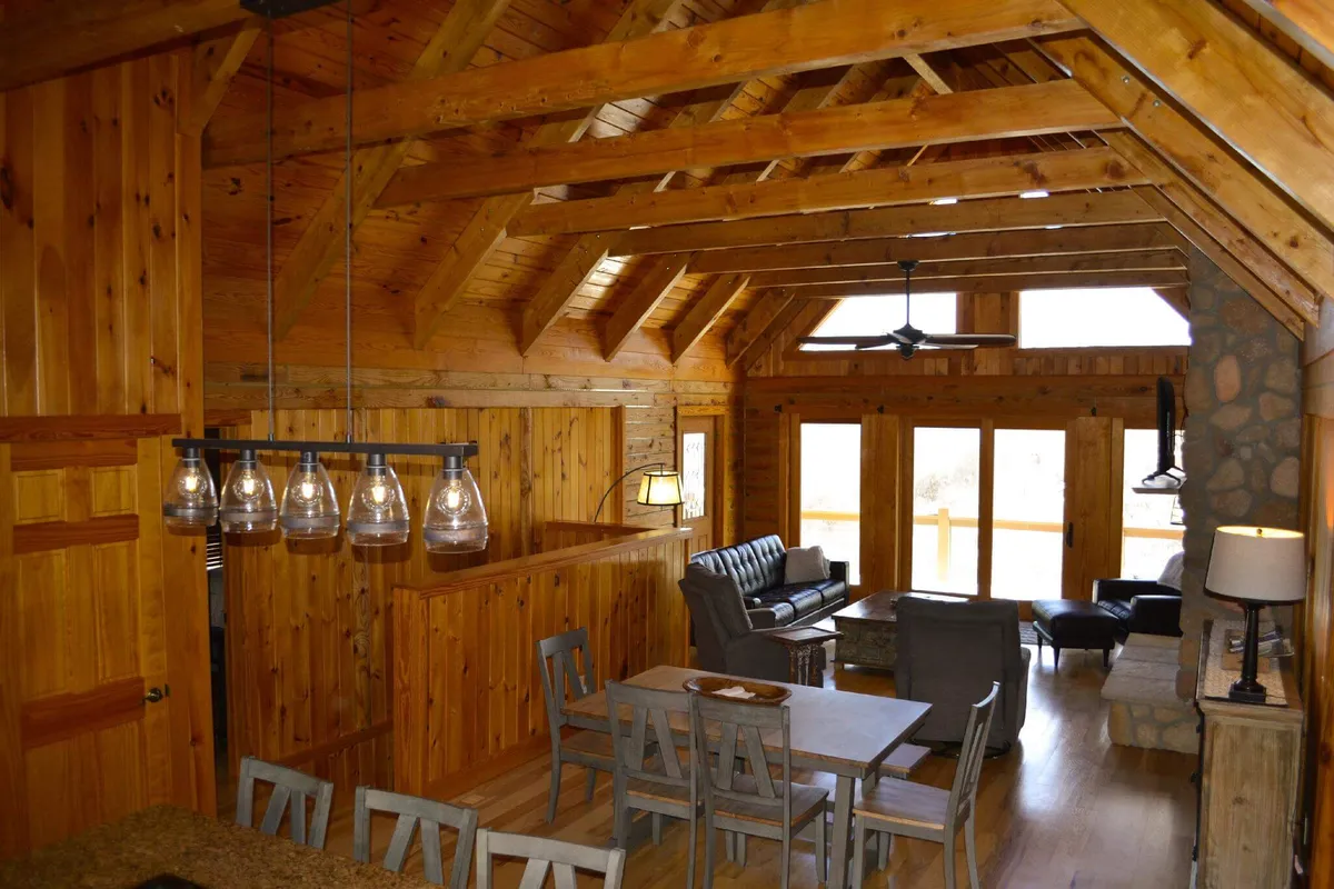 The image showcases a spacious, open-concept living and dining area with exposed wooden beams, a large dining table perfect for family meals, cozy sofa seating, and expansive windows that frame breathtaking views of the Blue Ridge Mountains. The warm, inviting atmosphere is accentuated by natural light flooding the space and the promise of memorable gatherings in a secluded mountain retreat.
