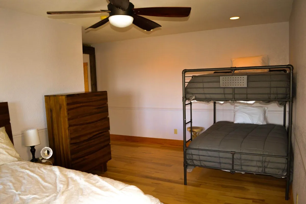 This interior scene captures a cozy guest room with a bunk bed, dresser, ceiling fan, and a nightstand with a lamp. The space feels inviting and versatile, suitable for kids, teens, or extra guests seeking a hideaway. The relaxed mood and practical furnishings echo the section's description of a flexible, tucked-away bedroom.