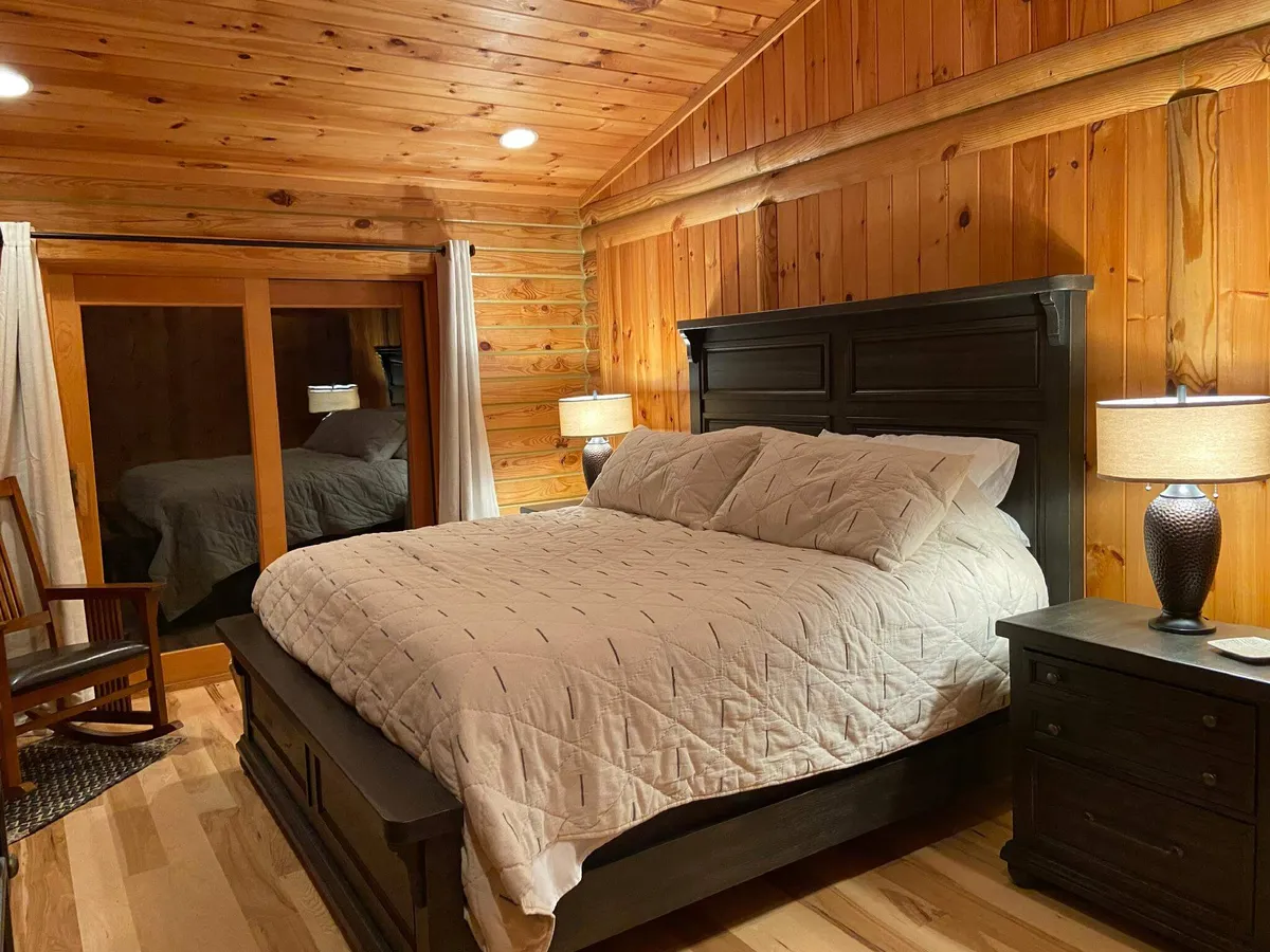 A warmly lit master bedroom featuring a king-size bed with luxury bedding, wooden walls, table lamps, and a cozy rocking chair. The mood is intimate and restful, perfect for a private retreat.
