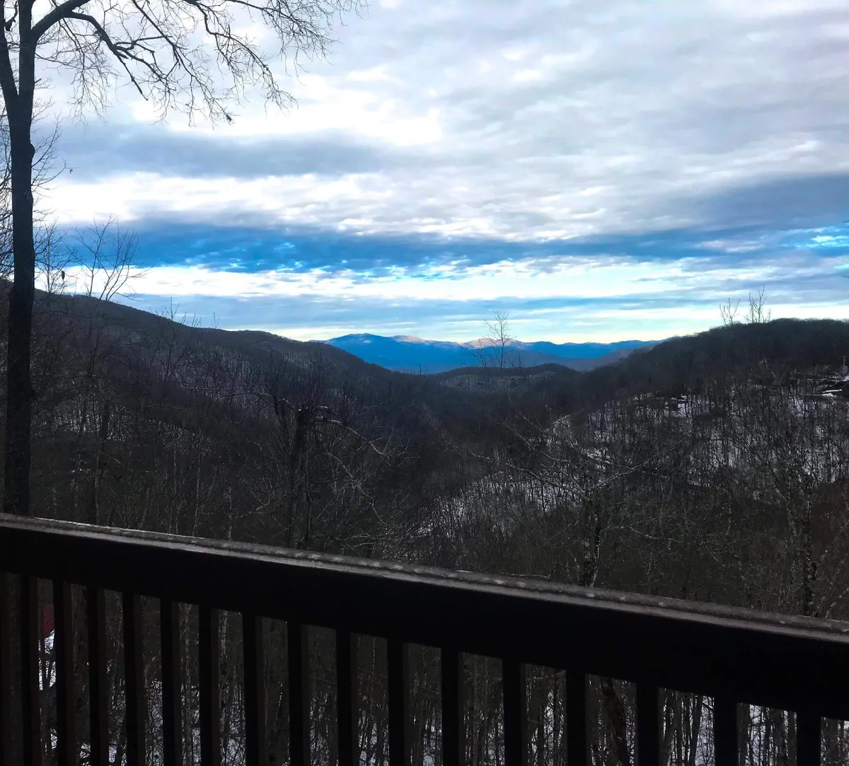 A panoramic view of a majestic mountain range under a cloudy sky with tree branches framing the scene in the foreground. The perspective evokes the feeling of being perched on a secluded deck, gazing out at nature's grandeur.