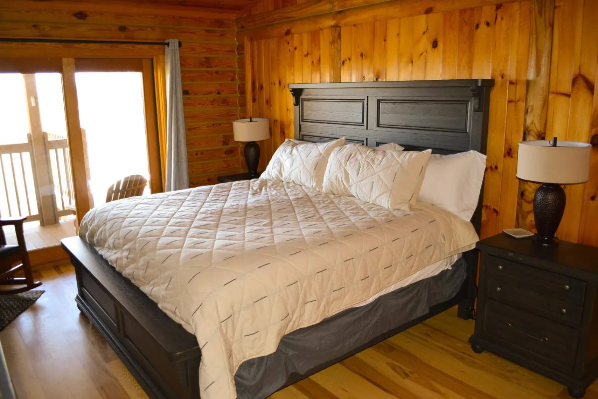 A welcoming bedroom with wooden walls, a comfortable bed, bedside lamps, and a rocking chair positioned near a window. Sunlight softly filters into the room, creating a calm and restful environment.