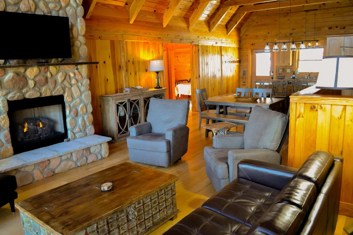 Another angle on the main gathering area, showcasing a stone fireplace, an inviting seating arrangement, a wooden dining table, and generous windows letting in the mountain landscape. The mood is relaxed and inviting, ideal for unwinding or socializing.
