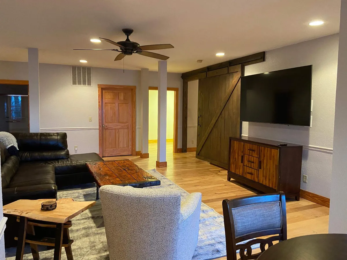 This interior shot centers on a warm, inviting entertainment room featuring a leather sectional sofa facing a large TV mounted on the wall. A wooden coffee table anchors the seating area, and a dining table is visible in the background. Notably, a custom barn door stands out, hinting at hidden spaces behind it. The overall mood is one of comfort and communal enjoyment, ideal for group gatherings or family movie nights.