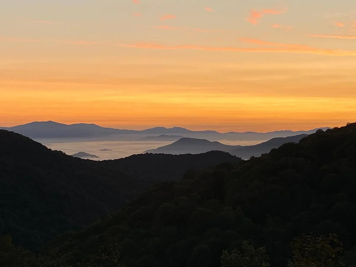 An expansive view of mountain silhouettes under vibrant sunset and twilight colors, with open sky and scattered clouds creating a dramatic and serene atmosphere. No immediate structures are visible, centering attention on the panoramic landscape.