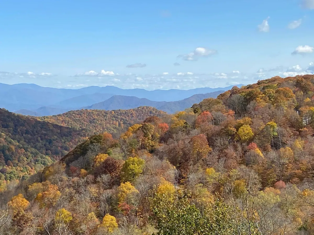 A breathtaking panoramic view of colorful autumn foliage blanketing distant mountain ranges beneath a blue sky with drifting clouds. The scene invites hiking, photography, and quiet appreciation of the natural landscape.